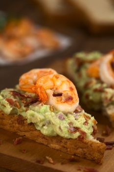 Wholegrain toast bread slices with guacamole, fried shrimp and fried bacon pieces on wooden board (Selective Focus, Focus on the front of the shrimp on the bread) 