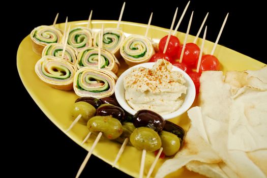 Plate of party food consisiting of olives, pickle onions, pita bread, hummus and ham and cheese roll ups.