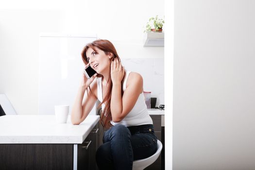 A young adult woman sitting in the kitchen while talking on a smartphone. 