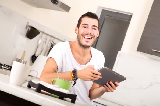 A young male sitting in the kitchen with a Tablet PC.