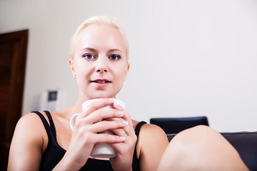 A blonde girl relaxing on a Sofa drinking a cup coffee
