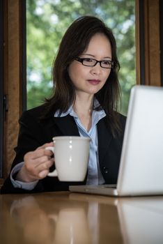 Woman in business suit with laptop on desk and picking up coffee in front of a window