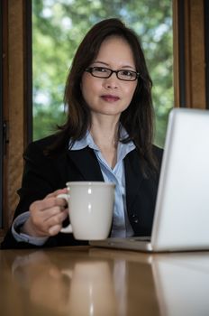 Woman in business suit with laptop on desk and picking up coffee in front of a window