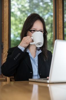 Woman in business suit with laptop on desk and drinking coffee in front of a window