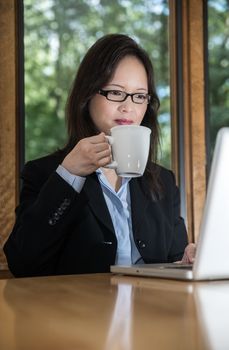 Woman in business suit with laptop on desk and drinking coffee in front of a window