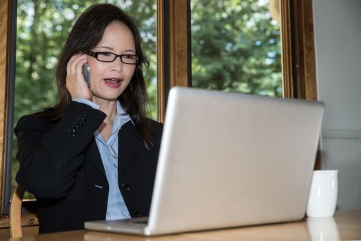 Woman in business suit with laptop and coffee on desk making a phone call in front of a window