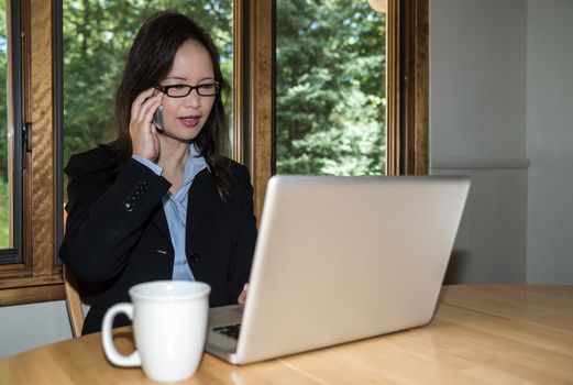 Woman in business suit with laptop and coffee on desk making a phone call in front of a window