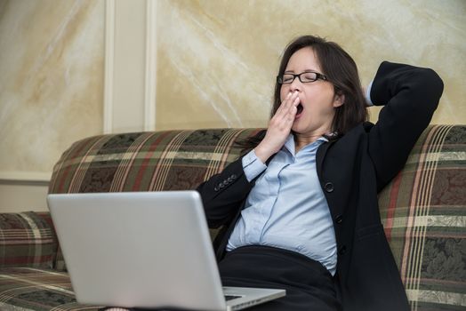 Woman in business suit feeling tired and yawning on sofa with labtop