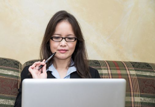 Professional woman in business suit on sofa in deep thought with pen and laptop 