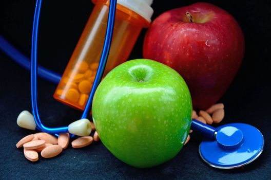 A red and green apple sits along side a stethoscope and pills