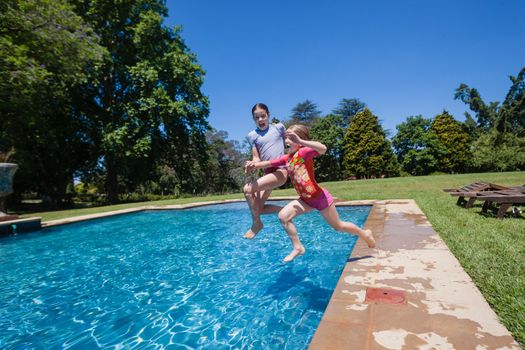 Young girls sisters having fun jumping into summer blue swimming pool.