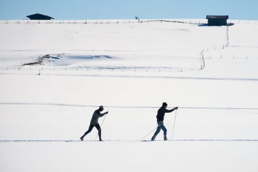 Two active cross country skiers following an x-country ski track across a flat expanse of winter snow exercising winter sports