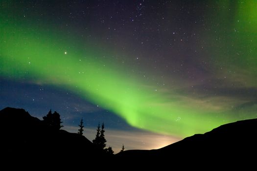 Intense bands of Northern lights or Aurora borealis or Polar lights dancing on night sky over hills with light glowing from moon rising behind them,Yukon Territory, Canada