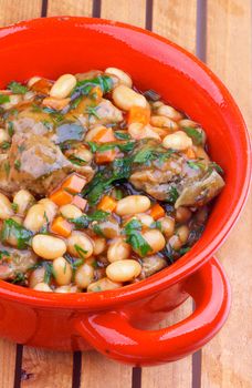 Homemade White Beans Stew with Boiled Beef and Greens in Rustic Red Pot closeup on Wooden background