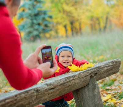 Photographing cute baby boy near bench in the autumn park