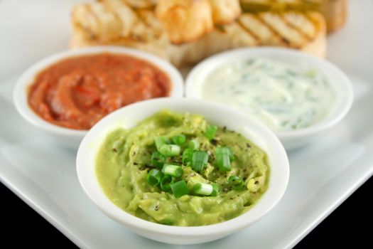 Three dips comprising guacamole, capsicum and tzatzki dips with bread.