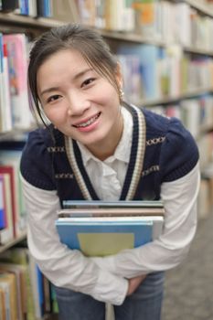 Portrait of young student holding a stack of text books near a library bookshelf