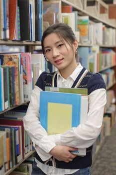 Portrait of young student holding a stack of text books near a library bookshelf