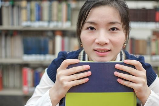 Portrait of clever girl student resting her chin on a text book in front of a library bookshelf