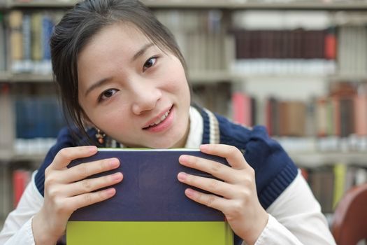 Portrait of clever girl student resting her chin on a text book in front of a library bookshelf