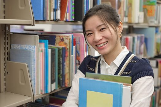 Portrait of young student holding a stack of text books near a library bookshelf