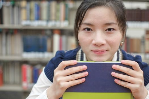 Portrait of clever girl student resting her chin on a text book in front of a library bookshelf