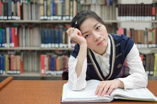 Young girl student with open textbook in front of a library bookshelf looking deep in thought