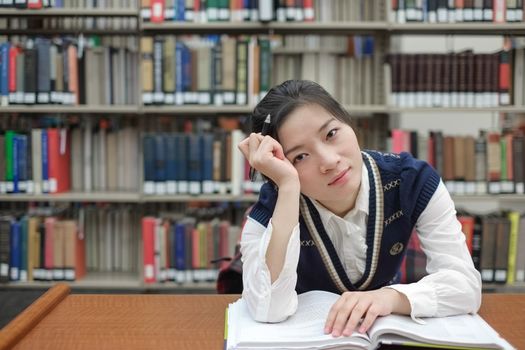 Young girl student with open textbook in front of a library bookshelf looking deep in thought