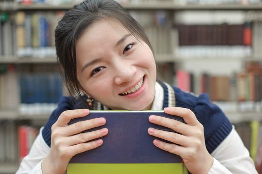 Portrait of clever girl student resting her chin on a text book in front of a library bookshelf