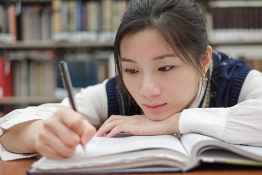 Tired young student resting her head on a textbook and doing homework in front of a library bookshelf