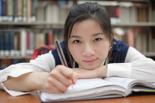 Tired young student resting her head on a textbook and doing homework in front of a library bookshelf