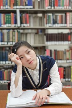 Young girl student with open textbook in front of a library bookshelf looking deep in thought