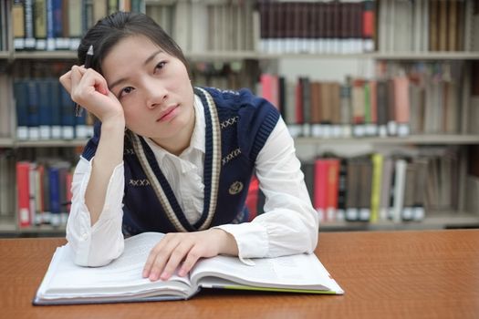 Young girl student with open textbook in front of a library bookshelf looking deep in thought