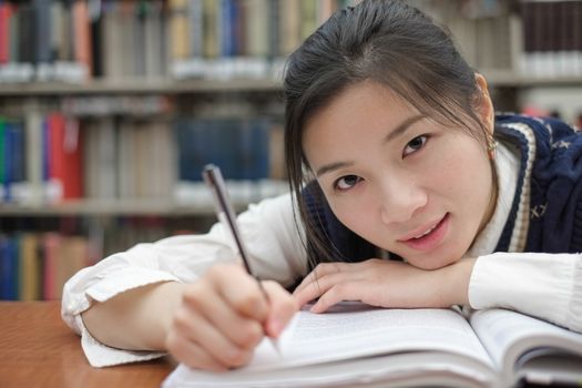 Tired young student resting her head on a textbook and doing homework in front of a library bookshelf