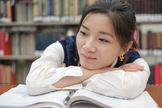 Tired young student resting her head on a textbook and doing homework in front of a library bookshelf