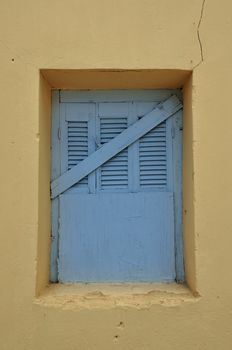 Blue window shutter and yellow cracked wall. Boarded up abandoned house exterior.