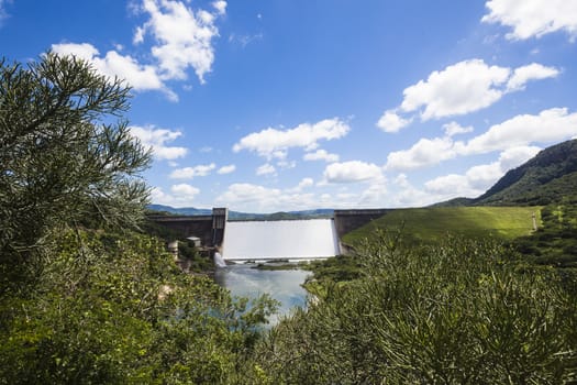 Dam filled to capacity and water flowing over the high wall in green summer valley.