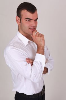 Handsome thoughtful, stylish man in a white shirt, smiling, with his arms crossed over his chest and his finger on the lip, side view on a gray background in studio