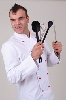 Happy handsome young male chef with a trendy hairstyle dressed in a white cooks jacket holding various kitchen utensils and laughing at the camera, studio portrait