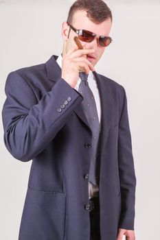 Suave successful businessman or CEO standing in a pair of sunglasses in his suit puffing on a big cigar