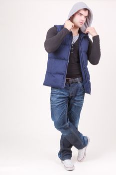 Relaxed charismatic young man in a hooded anorak top and jeans standing cross legged laughing at the camera, full length on white