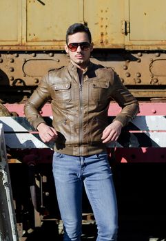 Attractive young man in leather jacket and jeans in front of old train wearing sunglasses