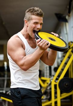 Handsome young man biting big weight disc in gym, looking at camera