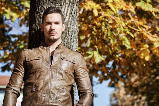 Handsome young man against tree in autumn, wearing leather jacket