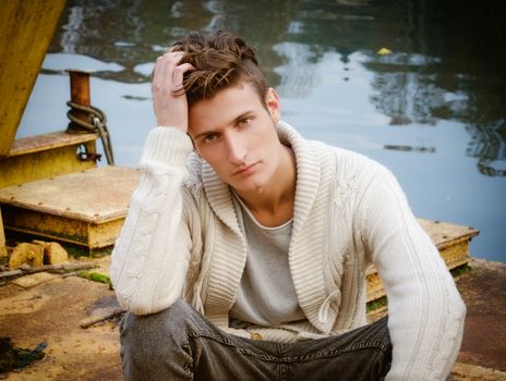 Attractive young man sitting on rusty metal next to river waters, looking at camera