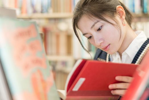 Beautiful female student reading a red covered book on a bookshelf in library