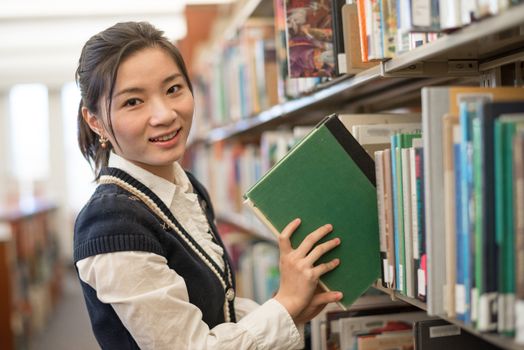 Young female student putting a green book back onto a bookshelf in library