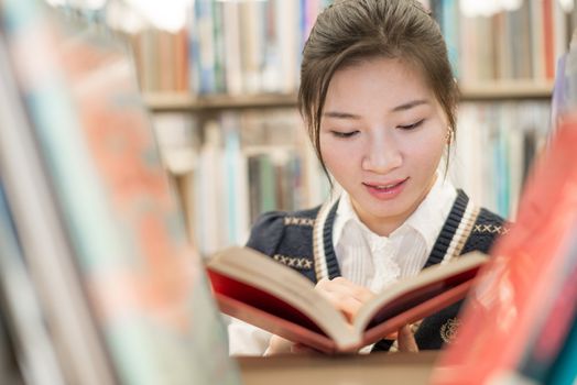 Beautiful female student reading a red covered book on a bookshelf in library