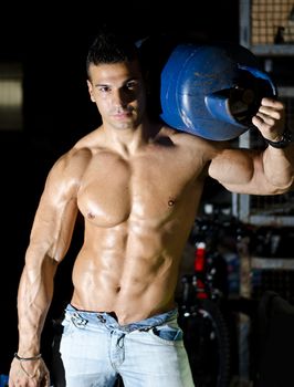 Muscular young man shirtless, carrying blue gas tank on shoulder, wearing only jeans