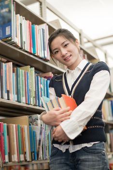 Portrait of cute young female student holding a stack of books next to a bookshelf in library